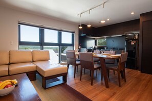 A modern kitchen with dark cabinetry, a dining area with a wooden table and chairs, and a large window providing a view of the outdoors. - Holiday Niseko Flow Niseko (Kutchan)