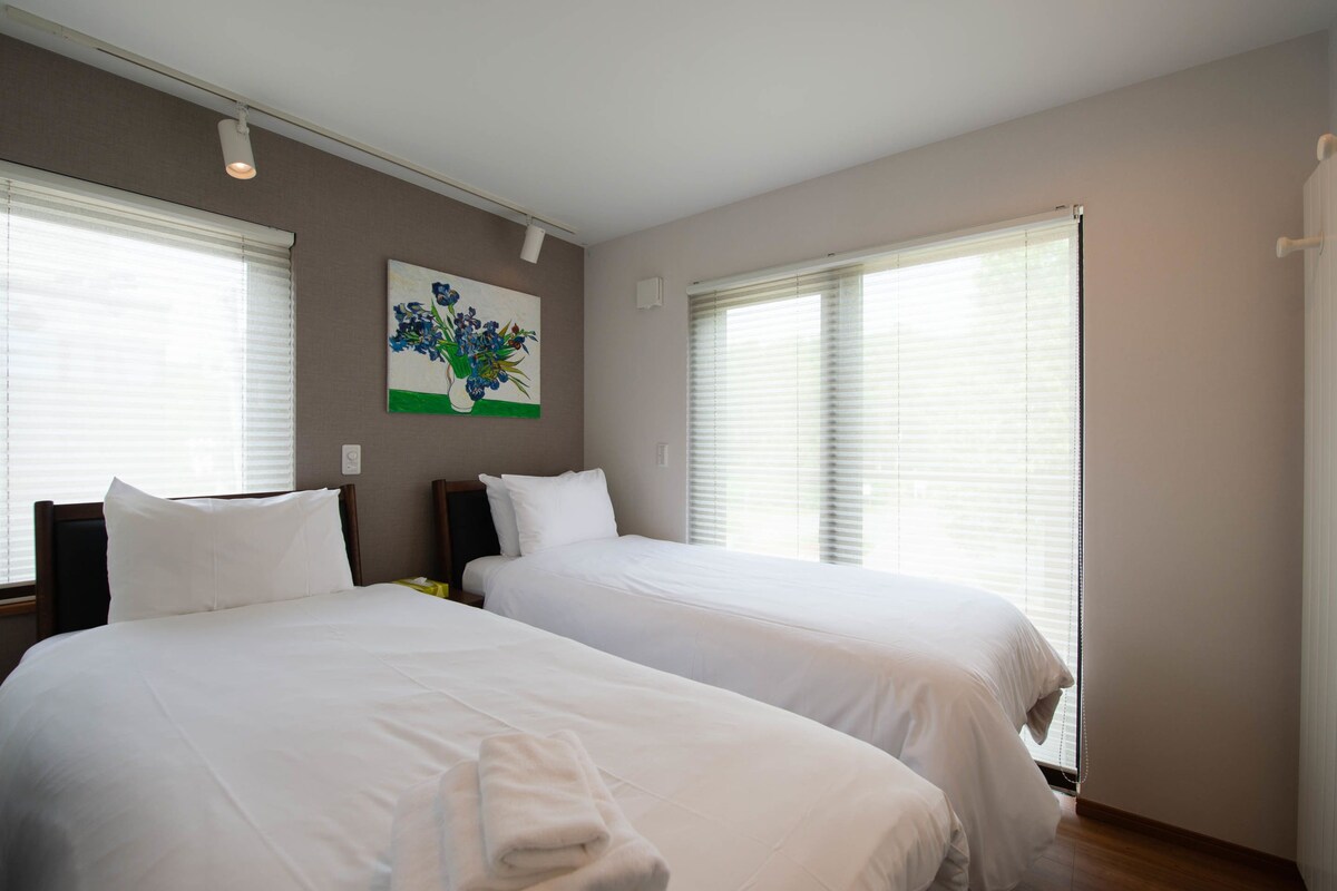 A hotel room with two beds, a painting on the wall, and large windows with blinds.