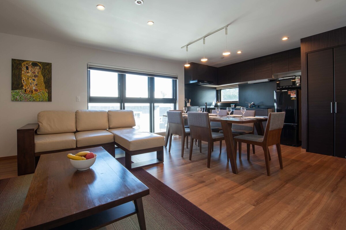 A modern living room with a sofa, dining table, and chairs. There is a kitchen area with cabinets and a refrigerator.