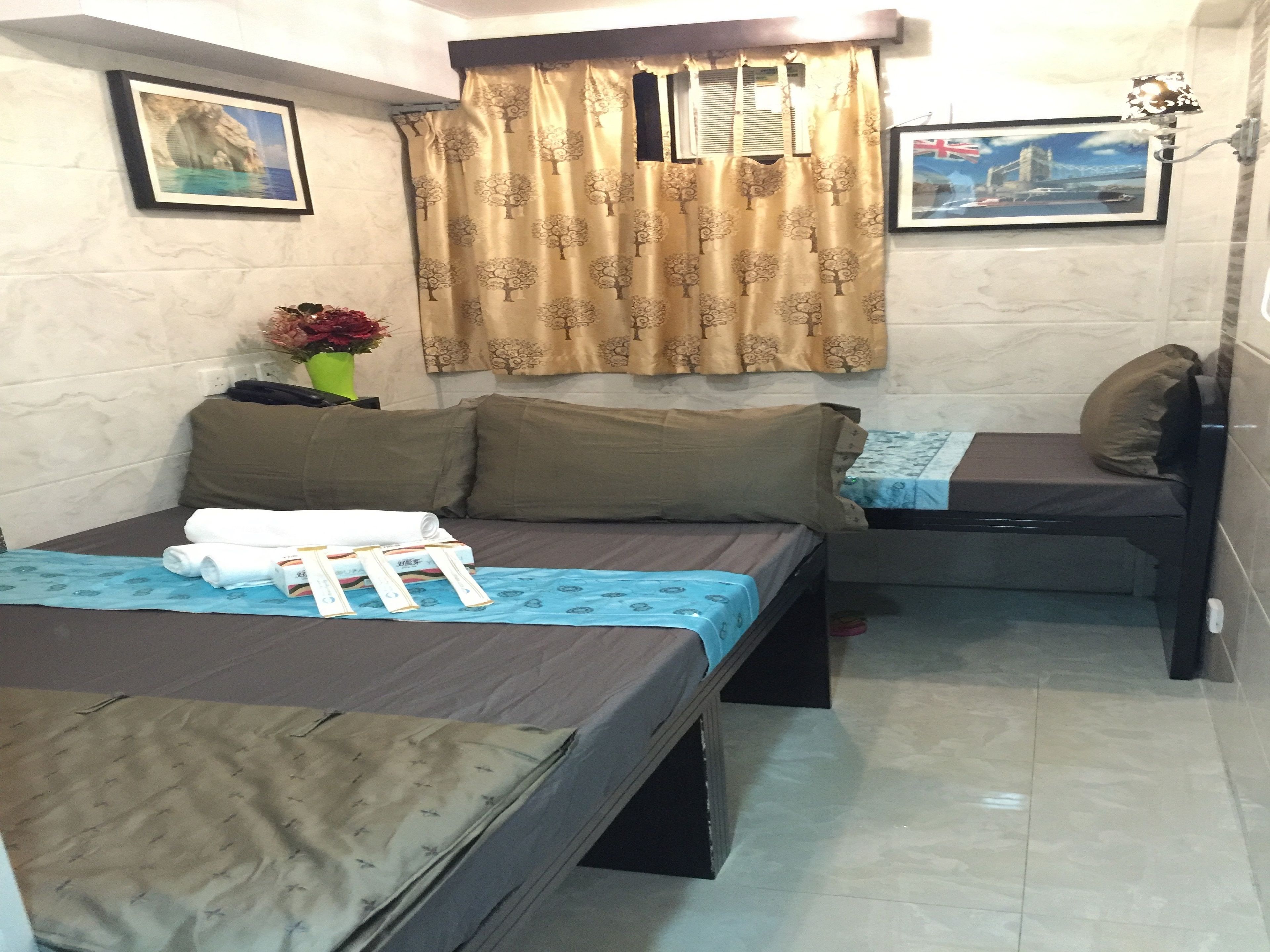 A small, compact room with two beds, a small table, and a window with curtains. There are pictures on the walls and a lamp on the table.