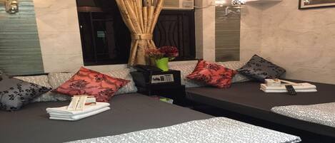 A hotel room with two beds, each with patterned bedding and pillows, a small table with a vase of flowers, and an air conditioning unit above the window.