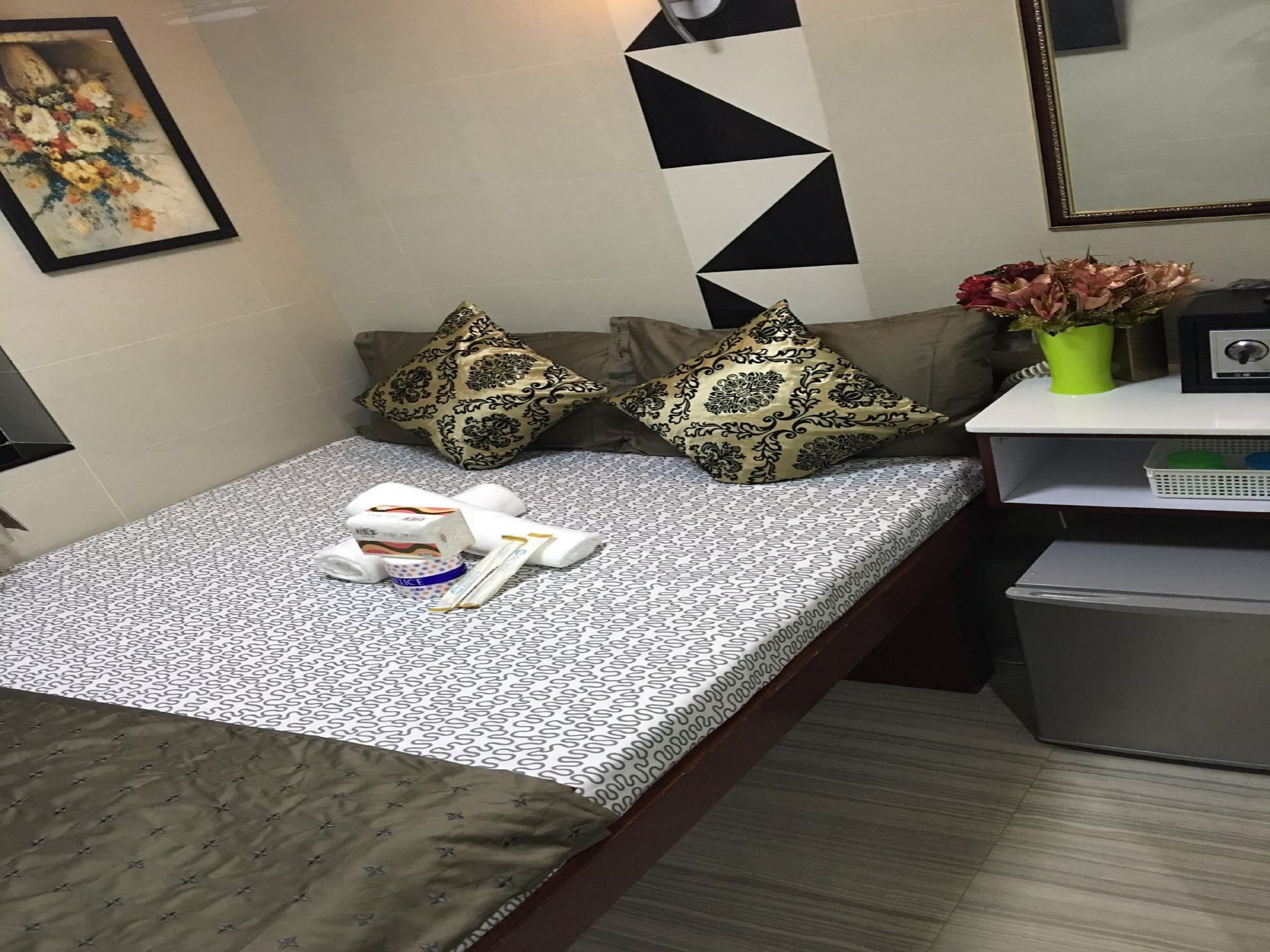A small, neatly arranged room with a bed, pillows, a bedside table with a radio, a vase with flowers, and a trash bin.