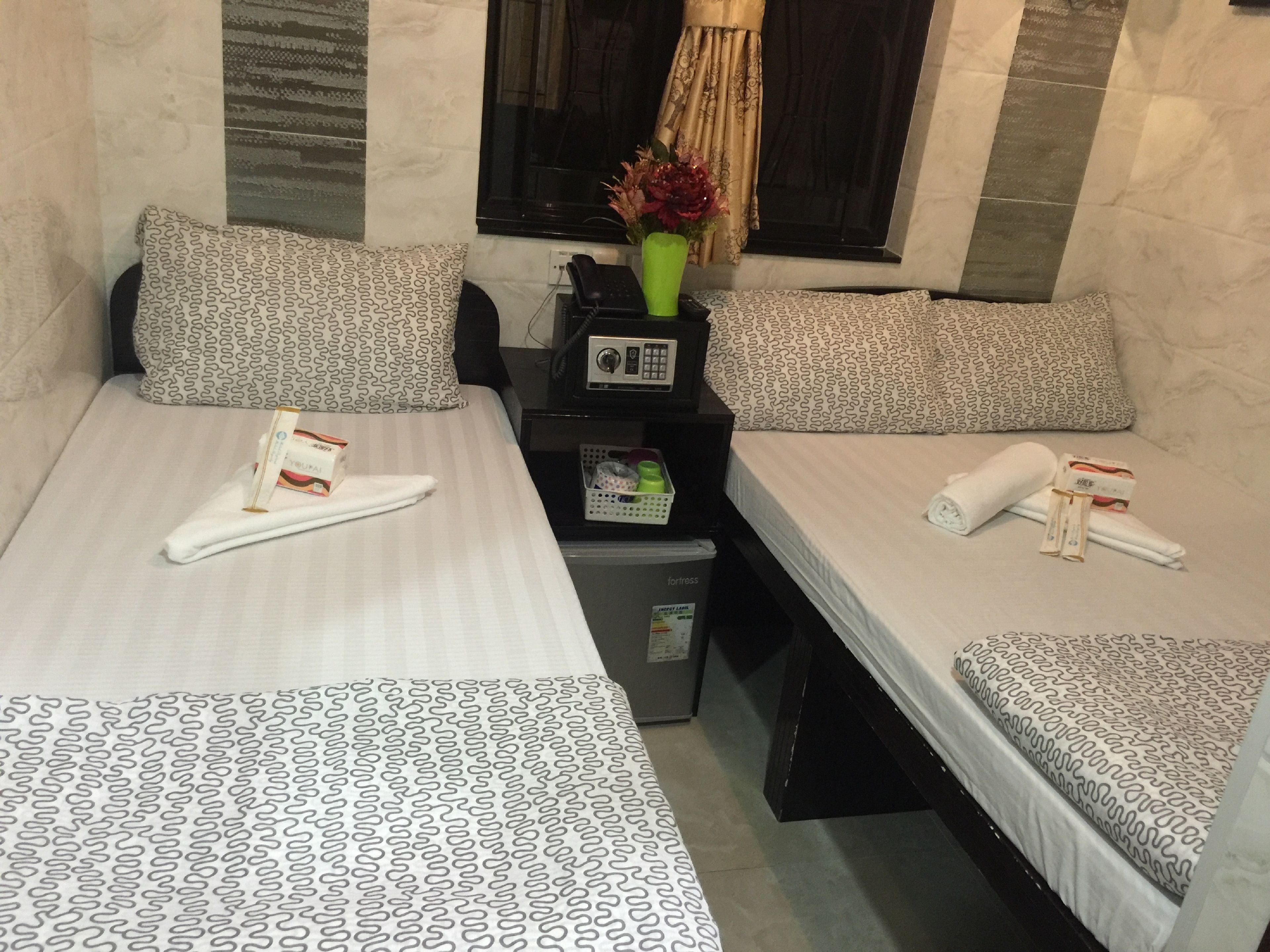 A hotel room with two beds, each with patterned bedding and pillows. There is a small table with a vase of flowers, a telephone, and toiletries.