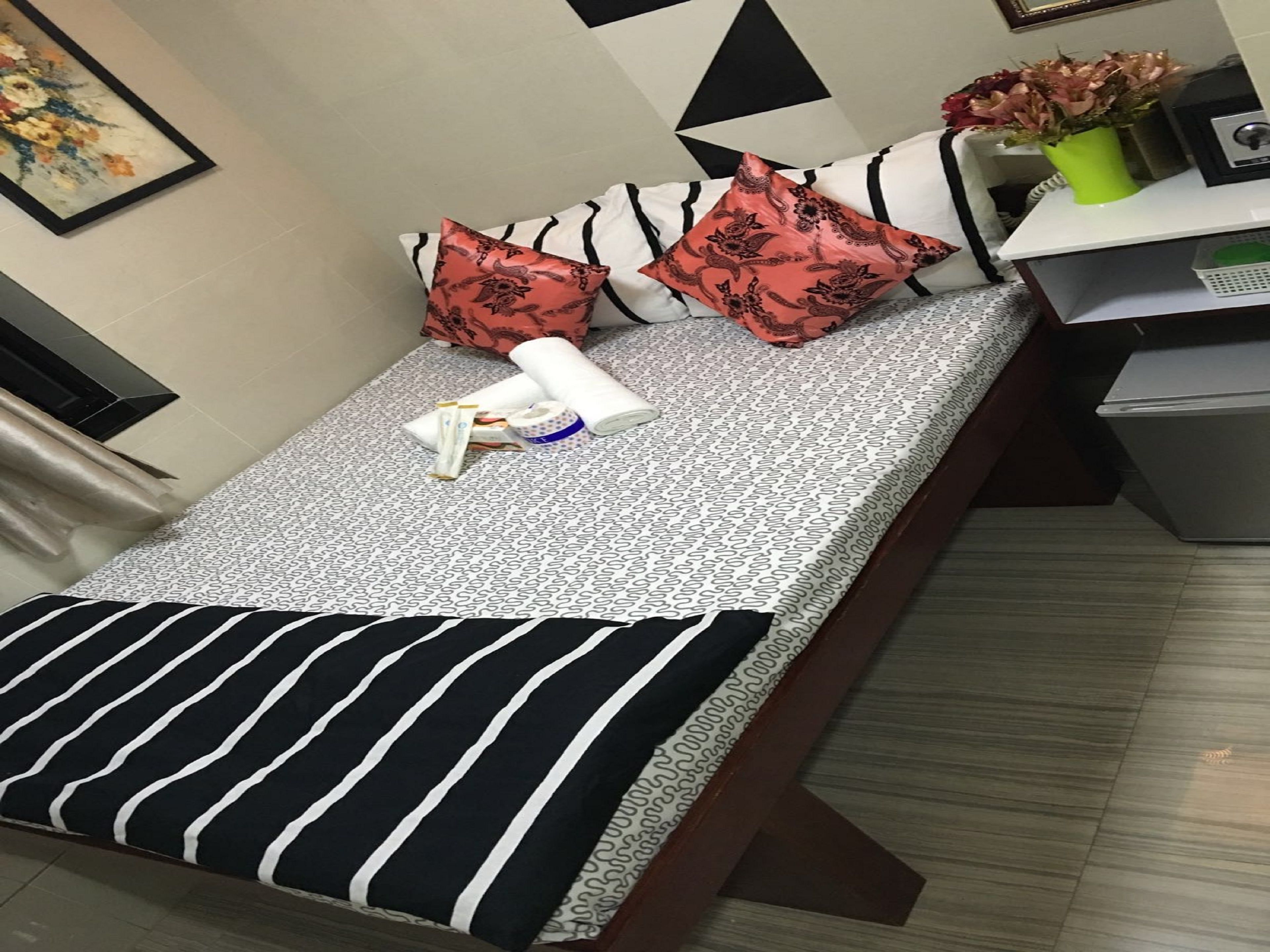 Una cama bien hecha con ropa de cama estampada, dos almohadas decorativas, una toalla blanca enrollada y una mesita de noche con un florero.