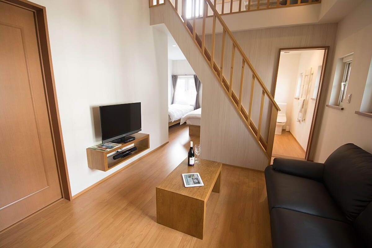 A modern living room with a wooden coffee table, a black leather sofa, a TV on a wooden shelf, and a staircase leading to another room.