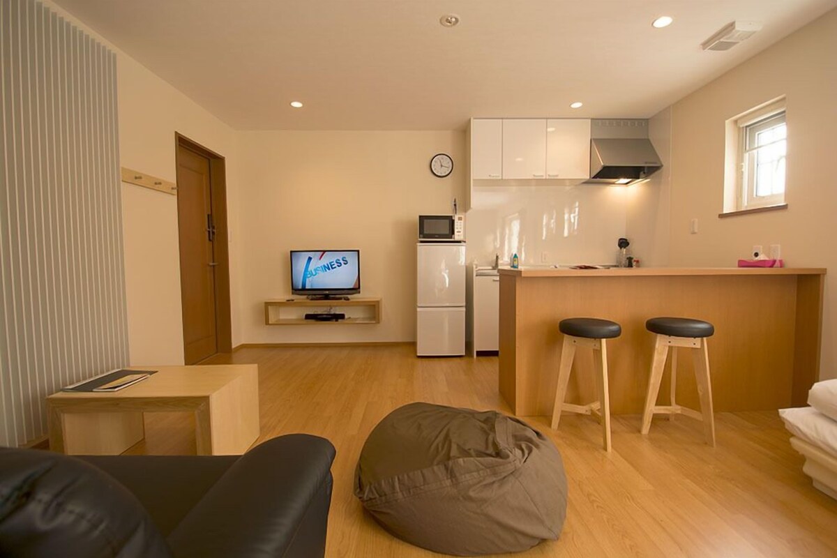 A compact living space with a kitchenette, a television, a clock, and a bean bag chair.