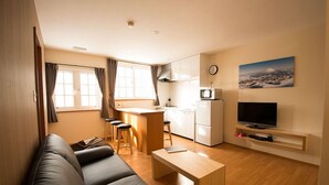 A compact living space with a sofa, a small dining area, a kitchen, a TV, and a painting of a mountain.