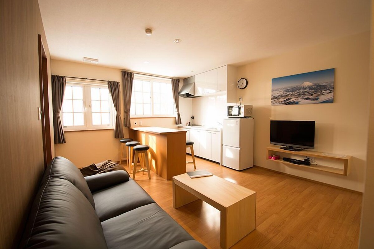 A compact living space with a sofa, a small dining area, a kitchen, a TV, and a painting of a mountain.