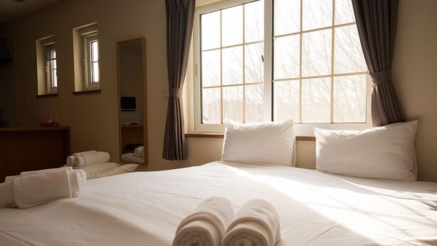 A hotel room with a large bed, two windows with curtains, a mirror, and a wooden side table with towels.