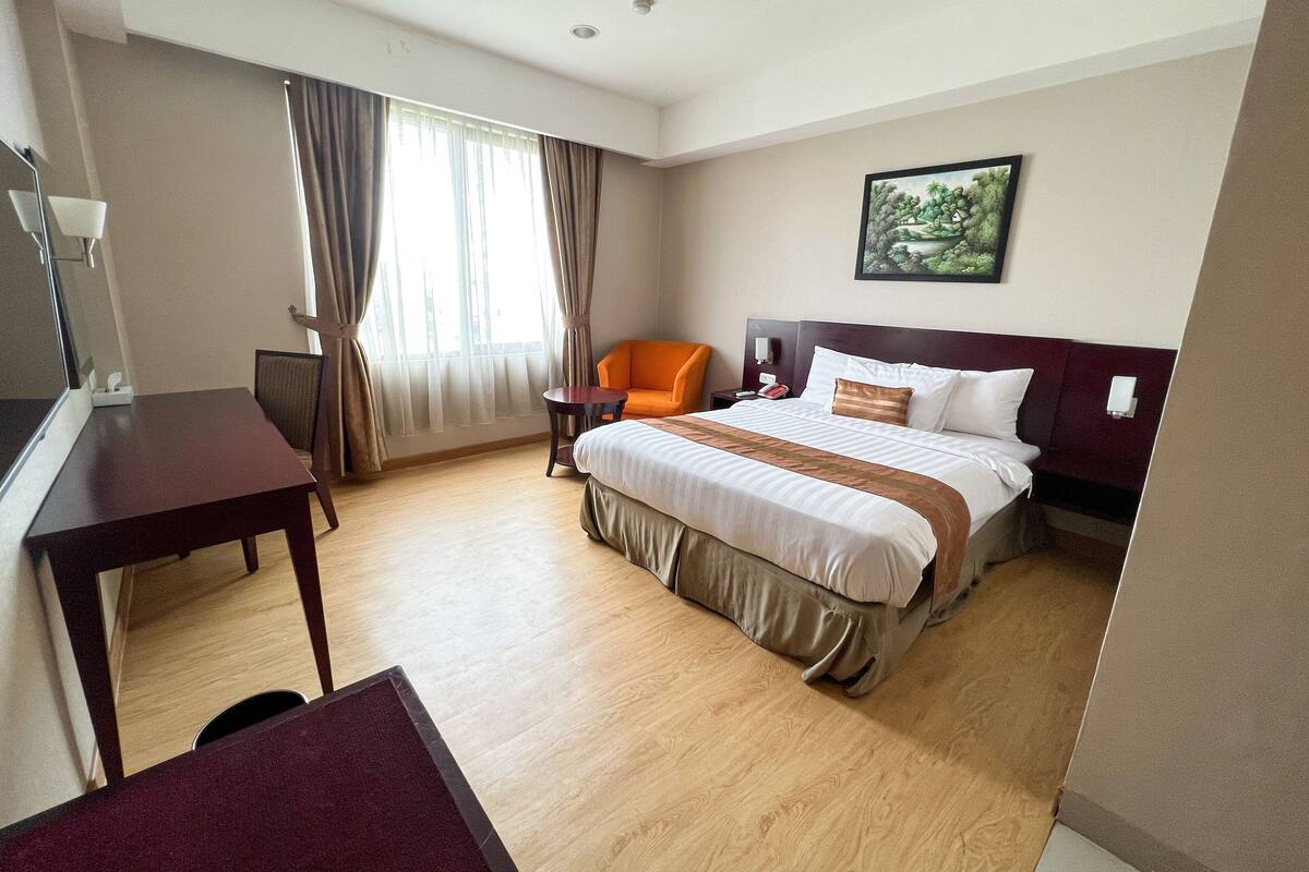 Une chambre d’hôtel avec un lit, un bureau, une chaise orange, une petite table et un tableau accroché au mur.