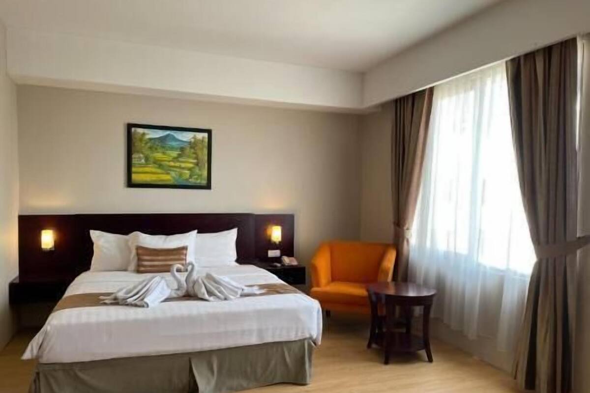 Une chambre d’hôtel avec un lit, une chaise, une petite table, un tableau au mur et une fenêtre avec des rideaux.
