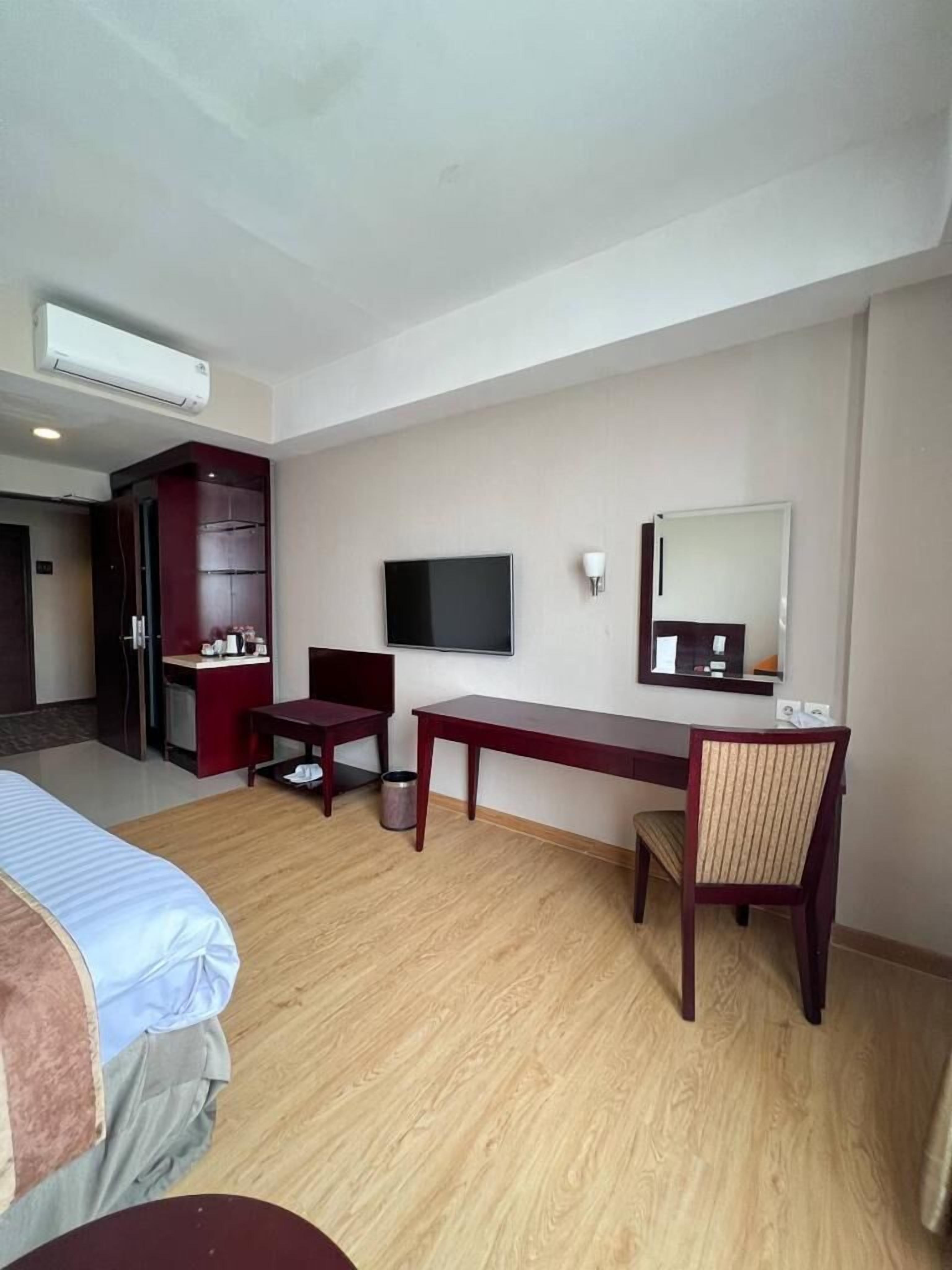 A hotel room with a bed, a desk, a chair, a television, a mirror, and an air conditioning unit.