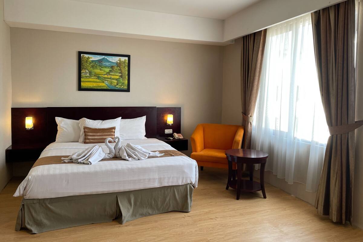 Une chambre d’hôtel avec un lit, une table de chevet, une chaise, une table, un tableau au mur et une fenêtre avec des rideaux.