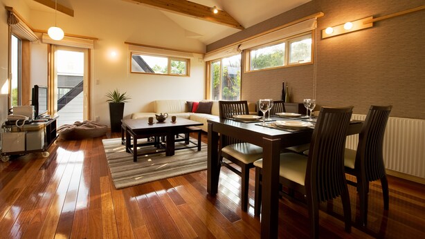 A spacious living area with a wooden floor, a sofa, a coffee table, and a dining table set for four. - Holiday Niseko Shinsetsu Apartments (Kutchan)