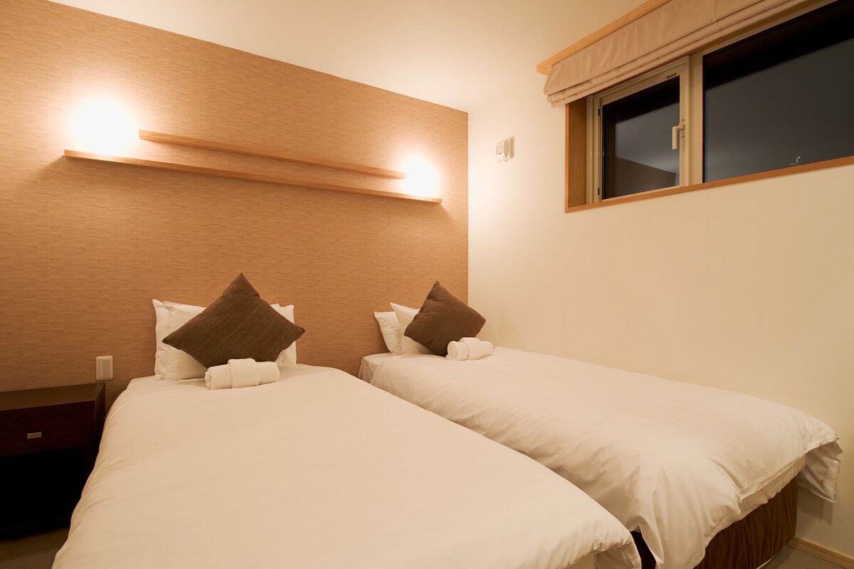 A hotel room with two single beds, a wooden headboard, bedside tables, and a window with a curtain.