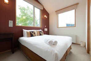 Habitación de hotel con una cama grande, cabecero de madera, dos ventanas con cortinas, una mesita de noche y un radiador.