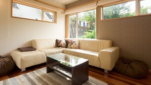 A modern living room with a beige sofa, a glass coffee table, and large windows offering a view of the outdoors. - Holiday Niseko Shinsetsu Apartments (Kutchan)