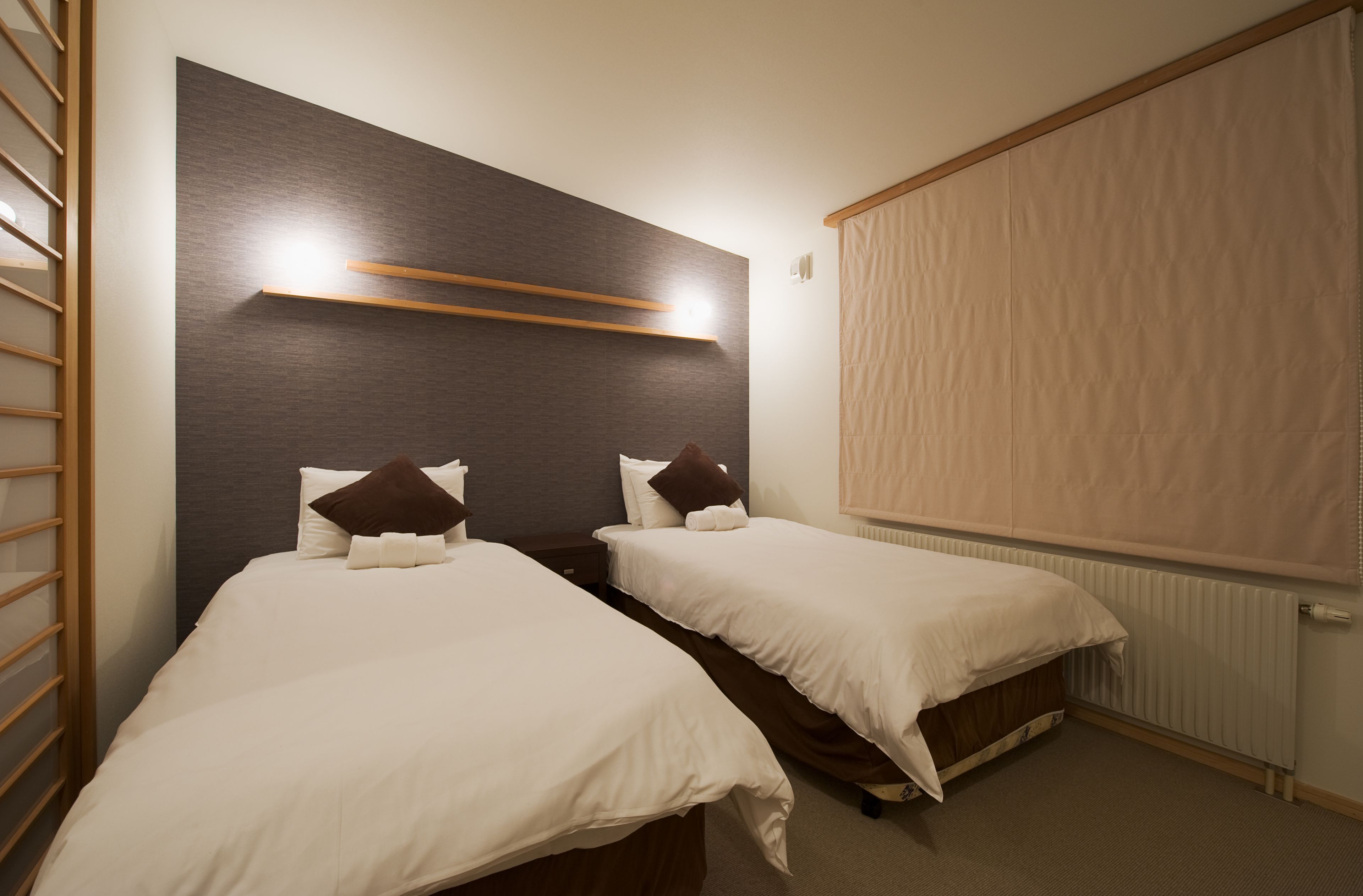 A room with two single beds, a wooden headboard, a window with blinds, and a radiator.