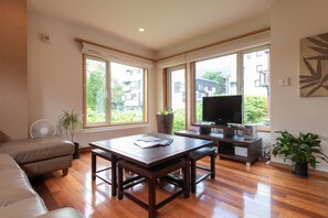 Un salón amplio con suelo de madera, un sofá, una mesa de comedor, un televisor y ventanas grandes que ofrecen vistas a edificios y zonas verdes.