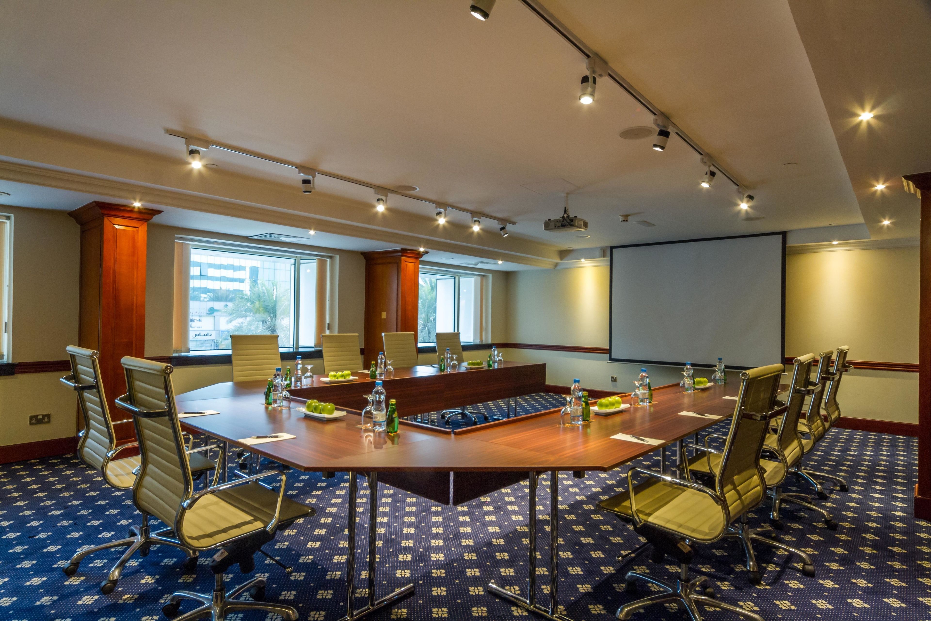 A conference room with a large oval table, multiple chairs, a projector screen, and a carpeted floor.