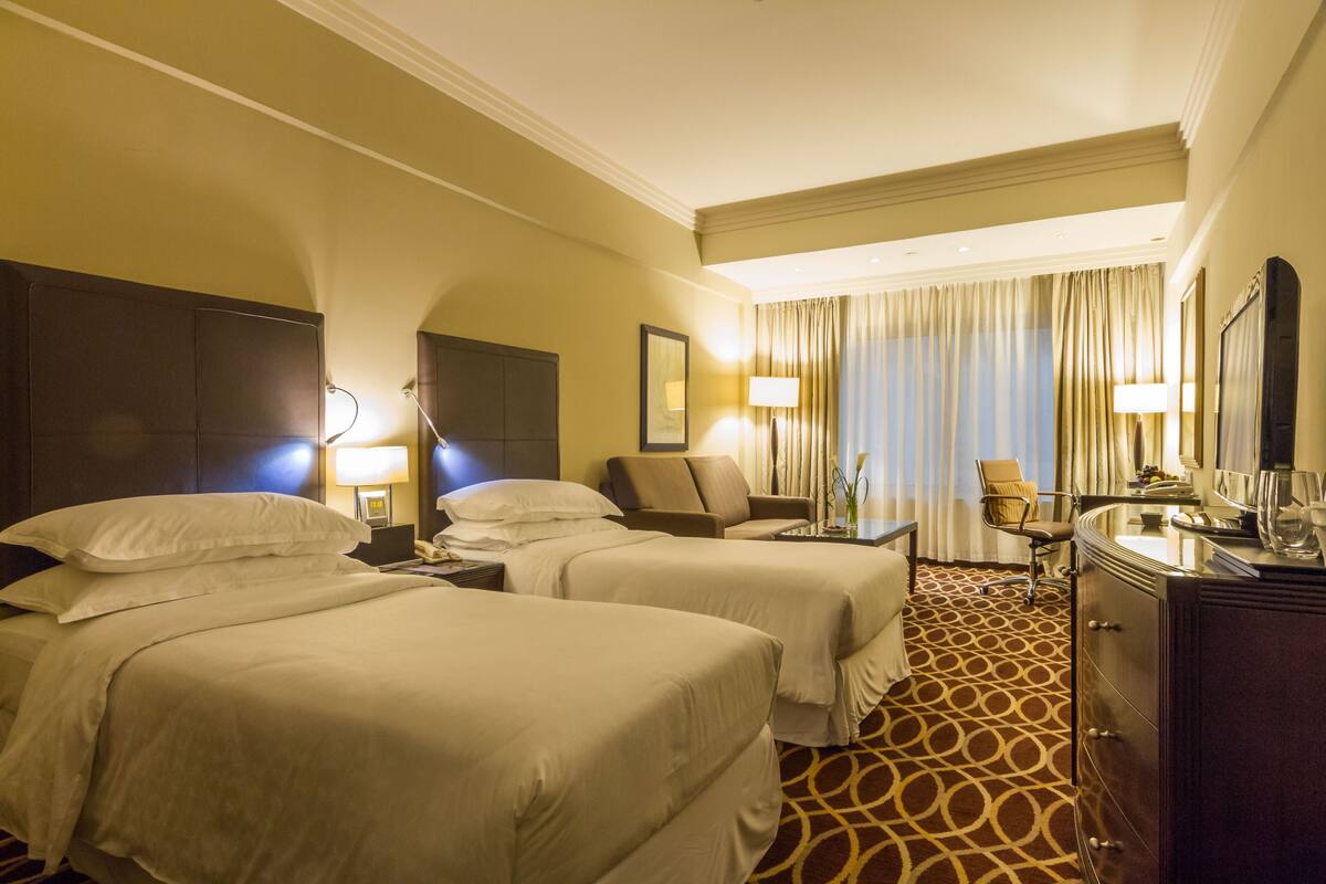 Ein Hotelzimmer mit zwei Betten, einem Schreibtisch, einem Stuhl, einem Schminktisch und einem großen Fenster mit Vorhängen.