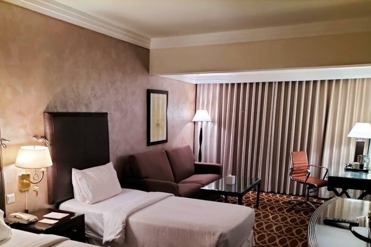 Ein Hotelzimmer mit Bett, Sofa, Schreibtisch und Stuhl. Auf dem Schreibtisch steht eine Lampe und an der Wand hängt ein gerahmtes Bild.