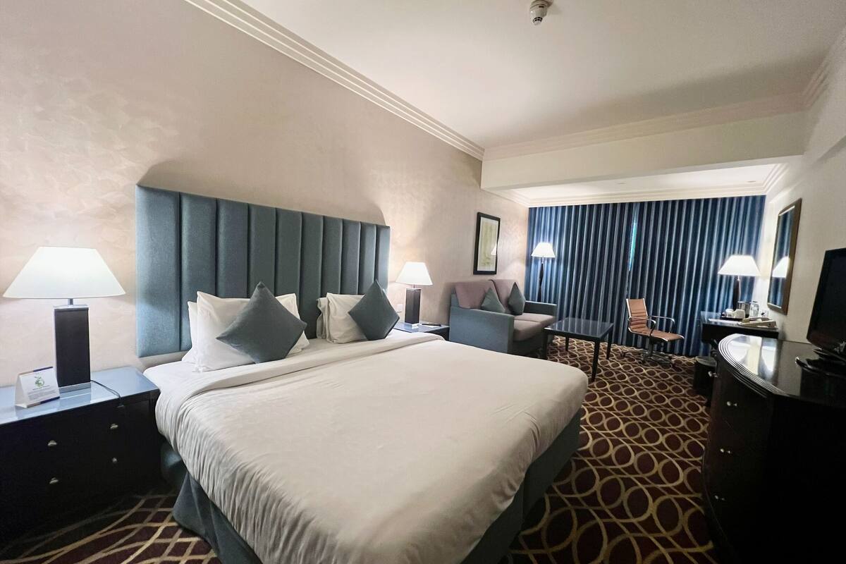 Ein Hotelzimmer mit einem großen Bett, zwei Nachttischen mit Lampen, einem Schreibtisch mit Stuhl, einem Fernseher und einem gemusterten Teppich.