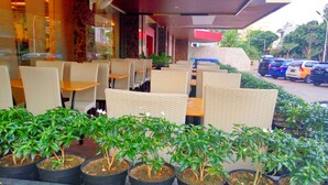 An outdoor seating area with wicker chairs and tables, potted plants, and a view of parked cars and greenery in the background. - The L Hotel Entertainment (Batam)