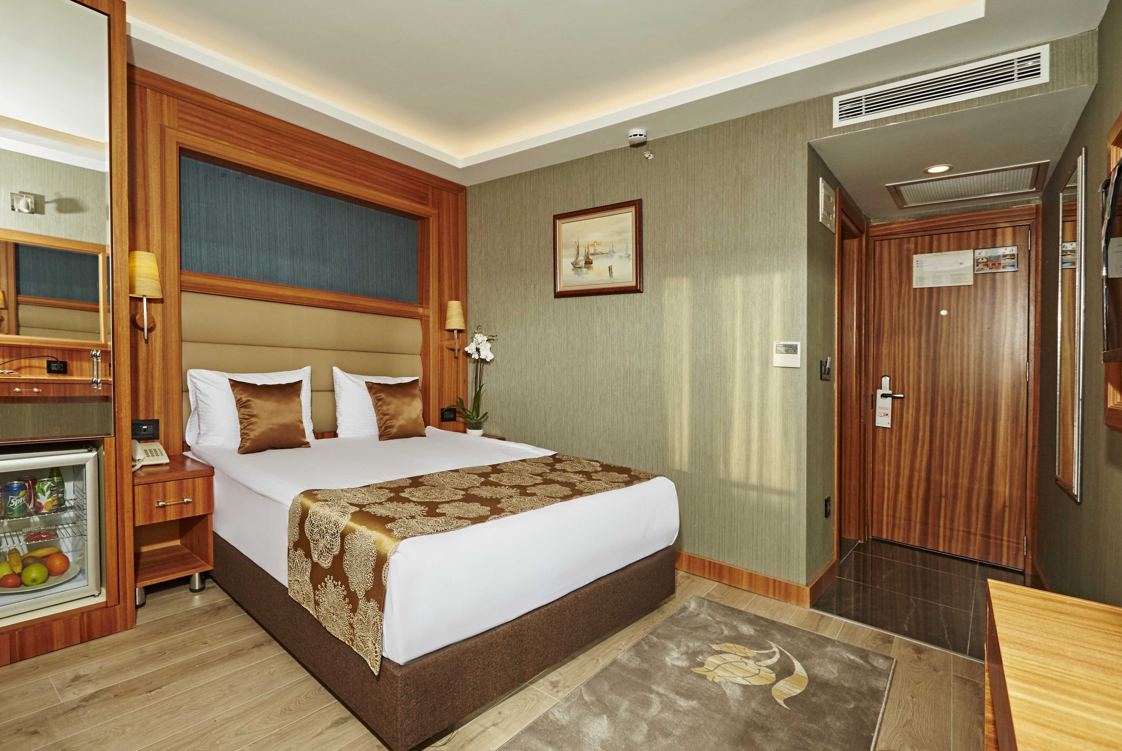 A hotel room with a large bed, a bedside table with a lamp, a small refrigerator, and a wooden wardrobe.