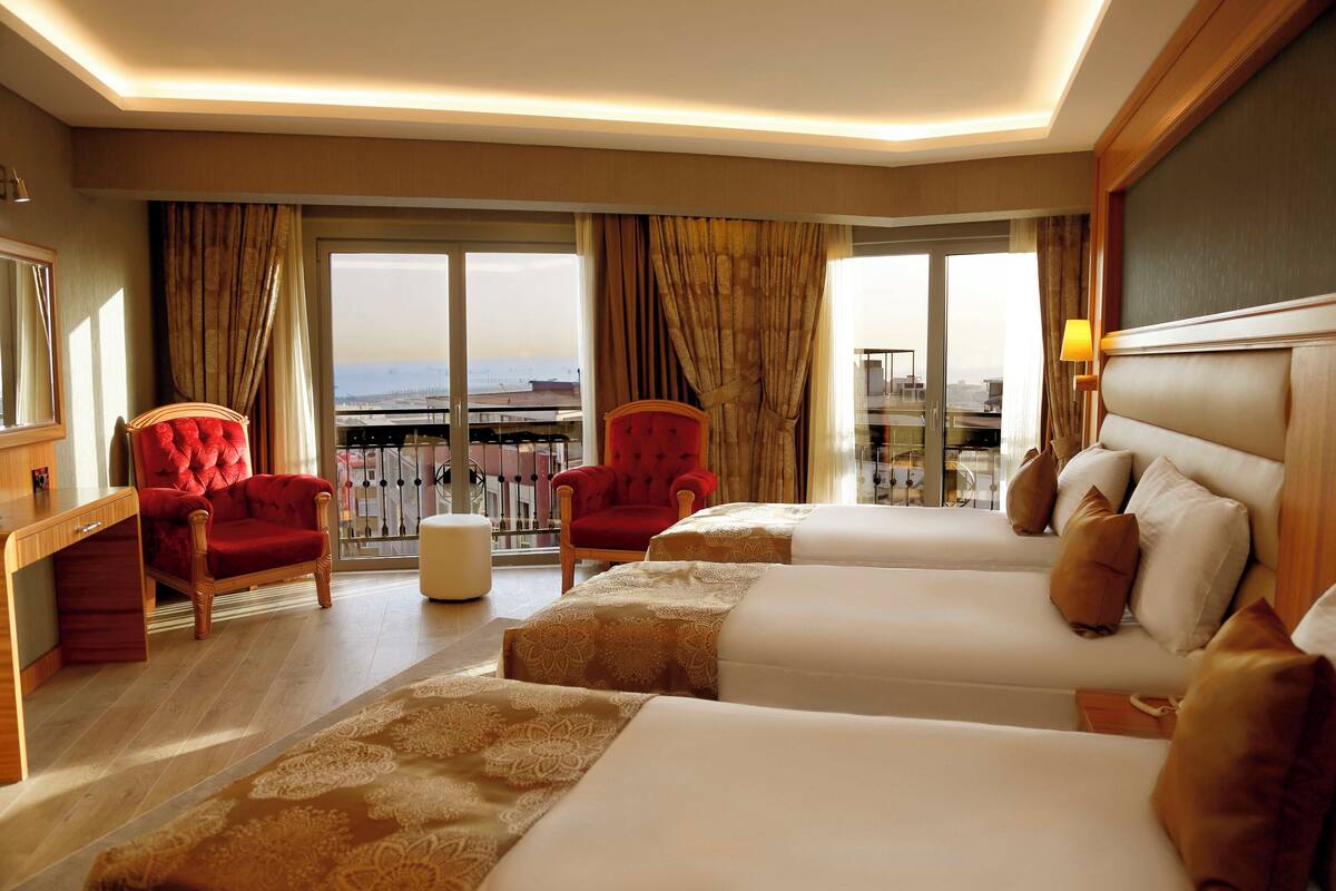 A hotel room with two beds, a red armchair, a wooden desk, and large windows offering a city view.