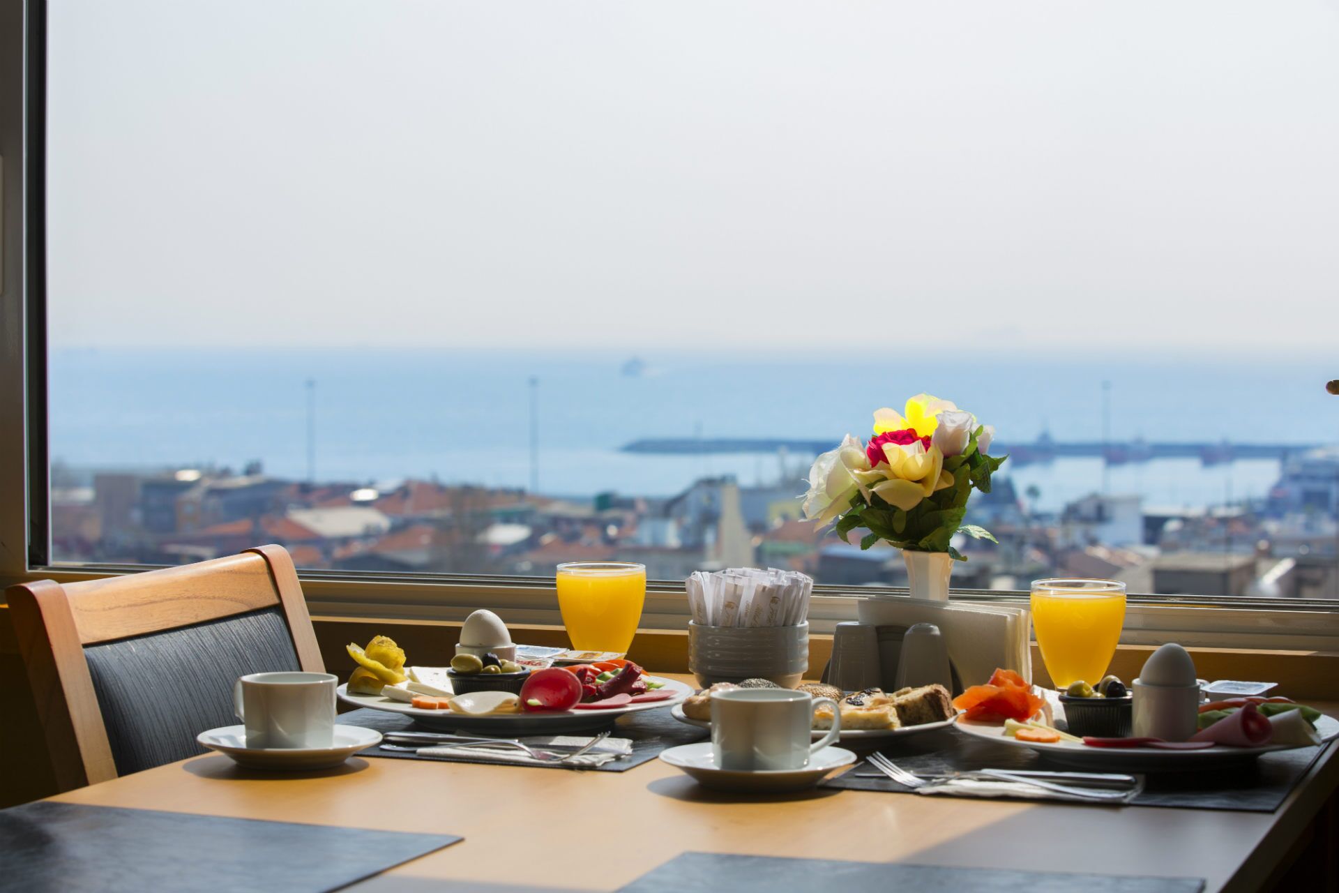 A breakfast table with a variety of food items and drinks, a vase with flowers, and a view of a coastal city through a window.