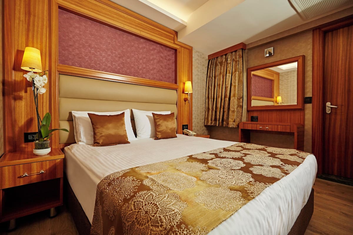 A hotel room with a large bed, wooden headboard, bedside lamps, a nightstand, a potted plant, a mirror, and a door with curtains.