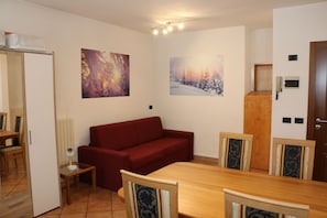 Una sala de estar con un sofá rojo, una mesa de comedor de madera y sillas, una mesita auxiliar y dos cuadros colgados en la pared.