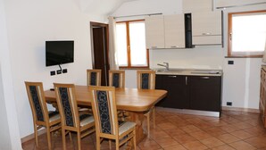 Un coin repas avec une table et des chaises en bois, un téléviseur fixé au mur et une cuisine équipée de placards et dotée d’une fenêtre.