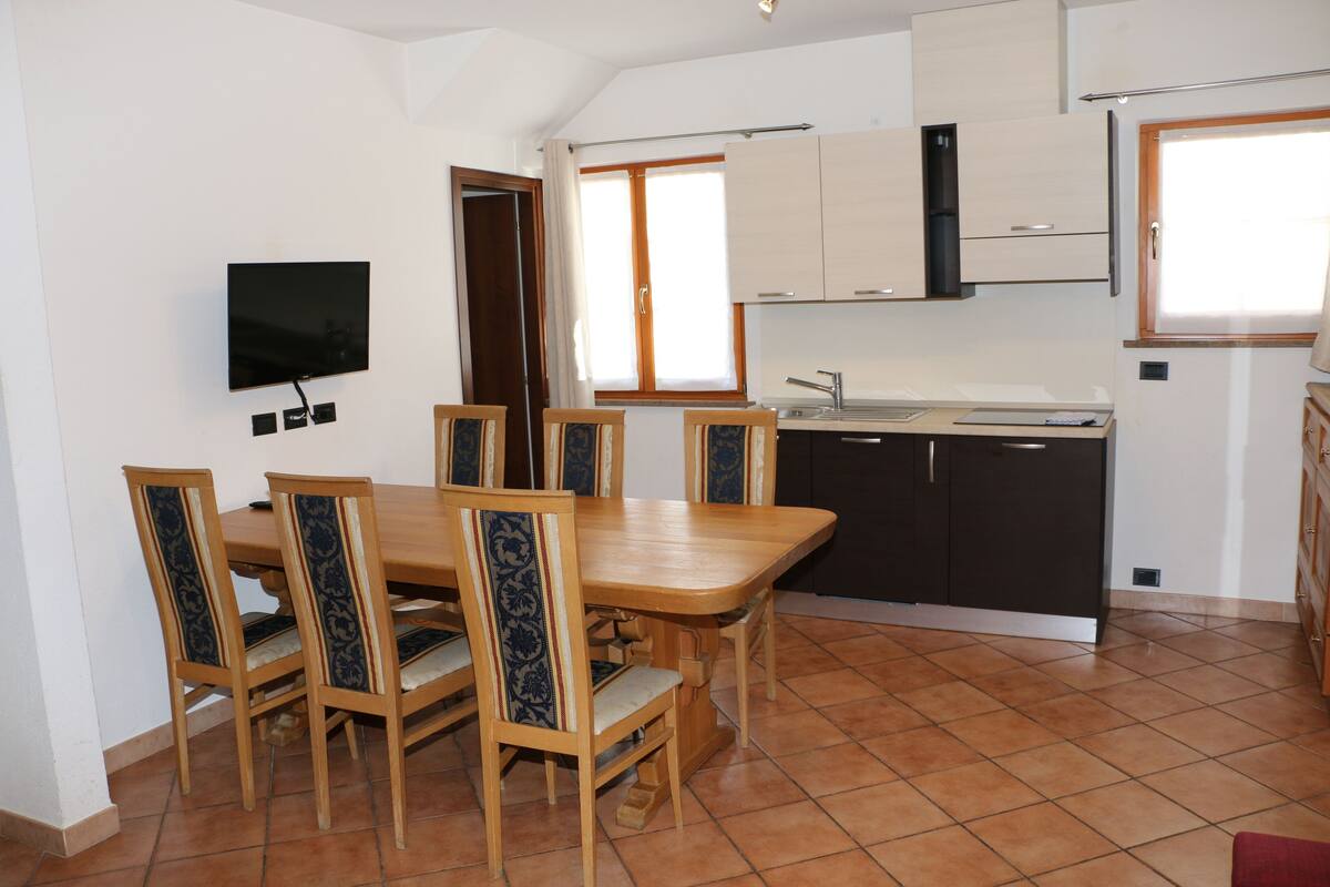 A dining area with a wooden table and chairs, a TV mounted on the wall, and a kitchen with cabinets and a window.