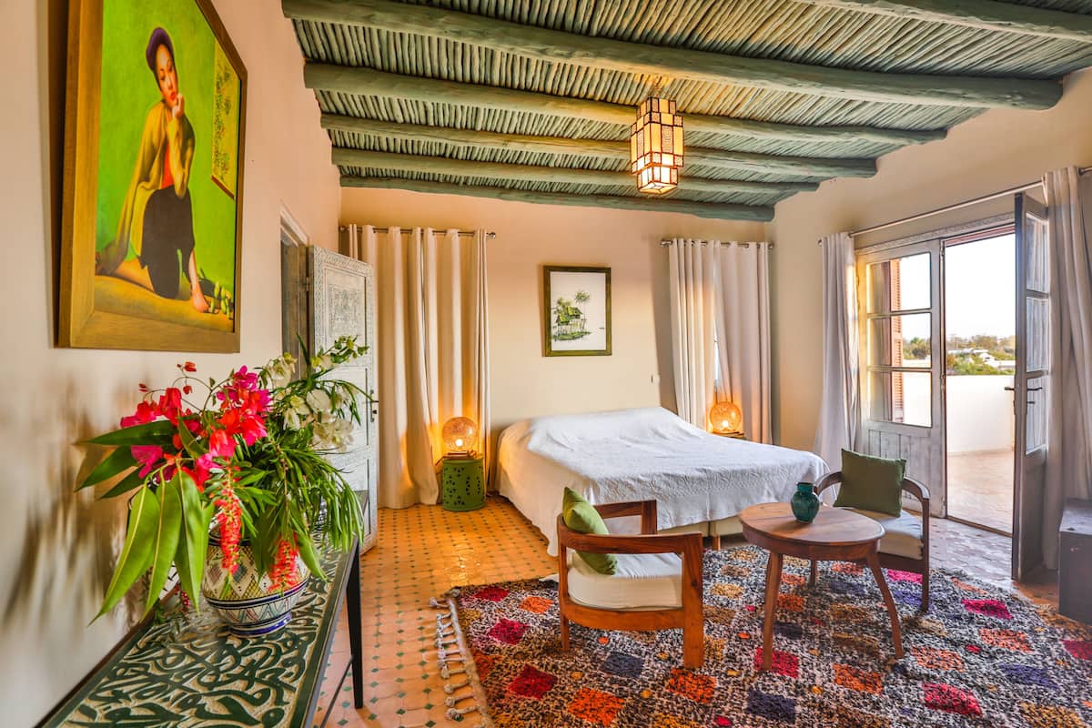 A bedroom with a bed, a chair, a small table, a vase with flowers, and a painting on the wall.