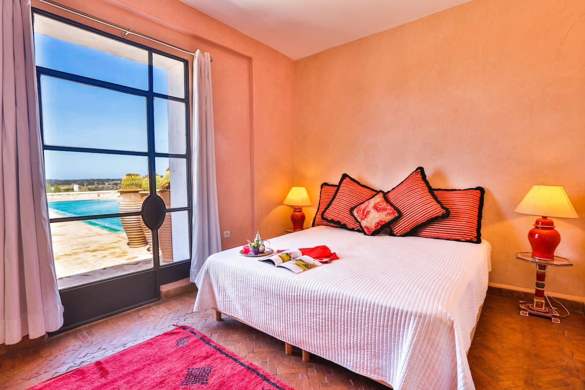 A bedroom with a large window, a bed with red and white bedding, two bedside lamps, and a red rug.