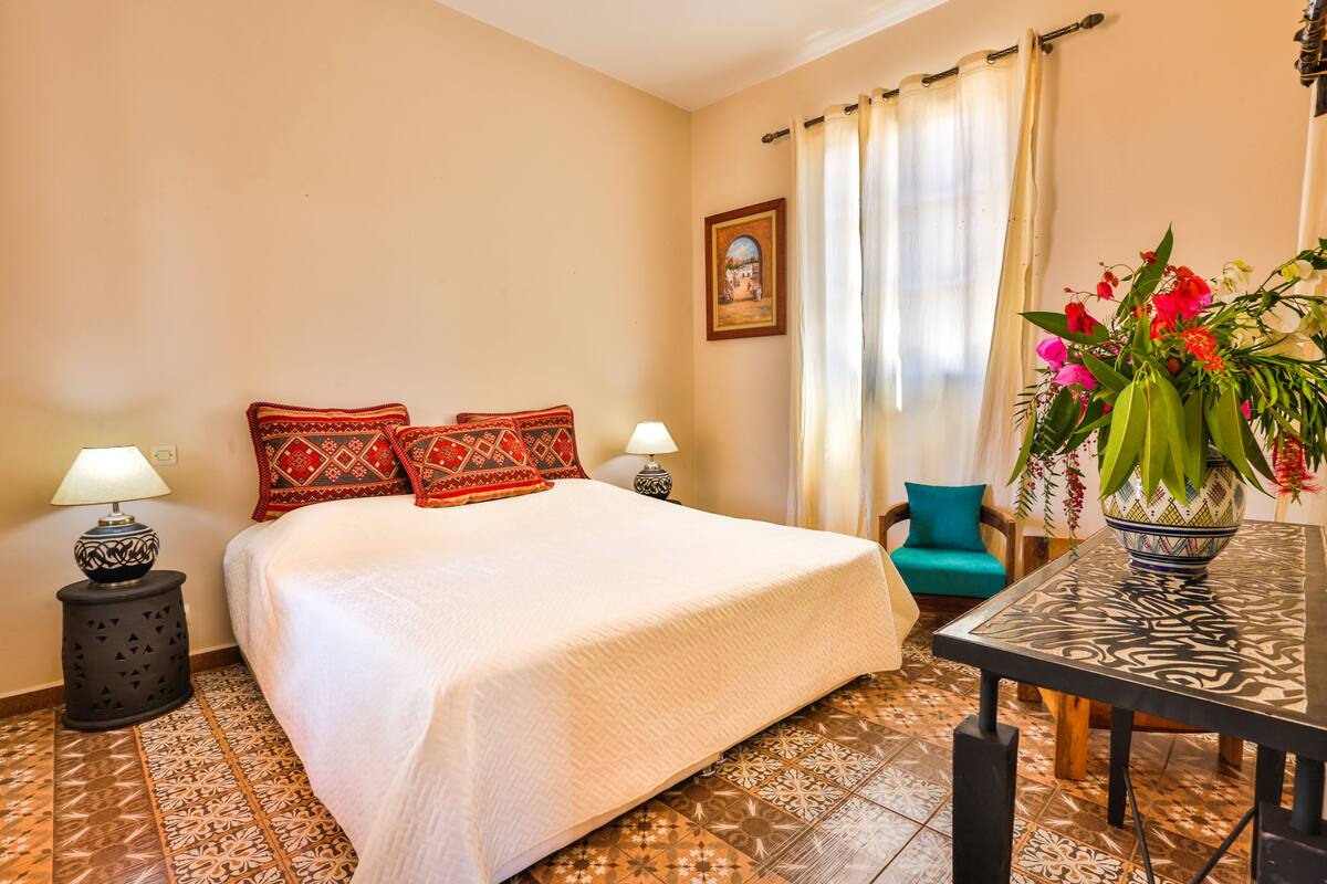 A neatly arranged bedroom with a bed, two bedside lamps, a chair, a table with a vase of flowers, and a painting on the wall.