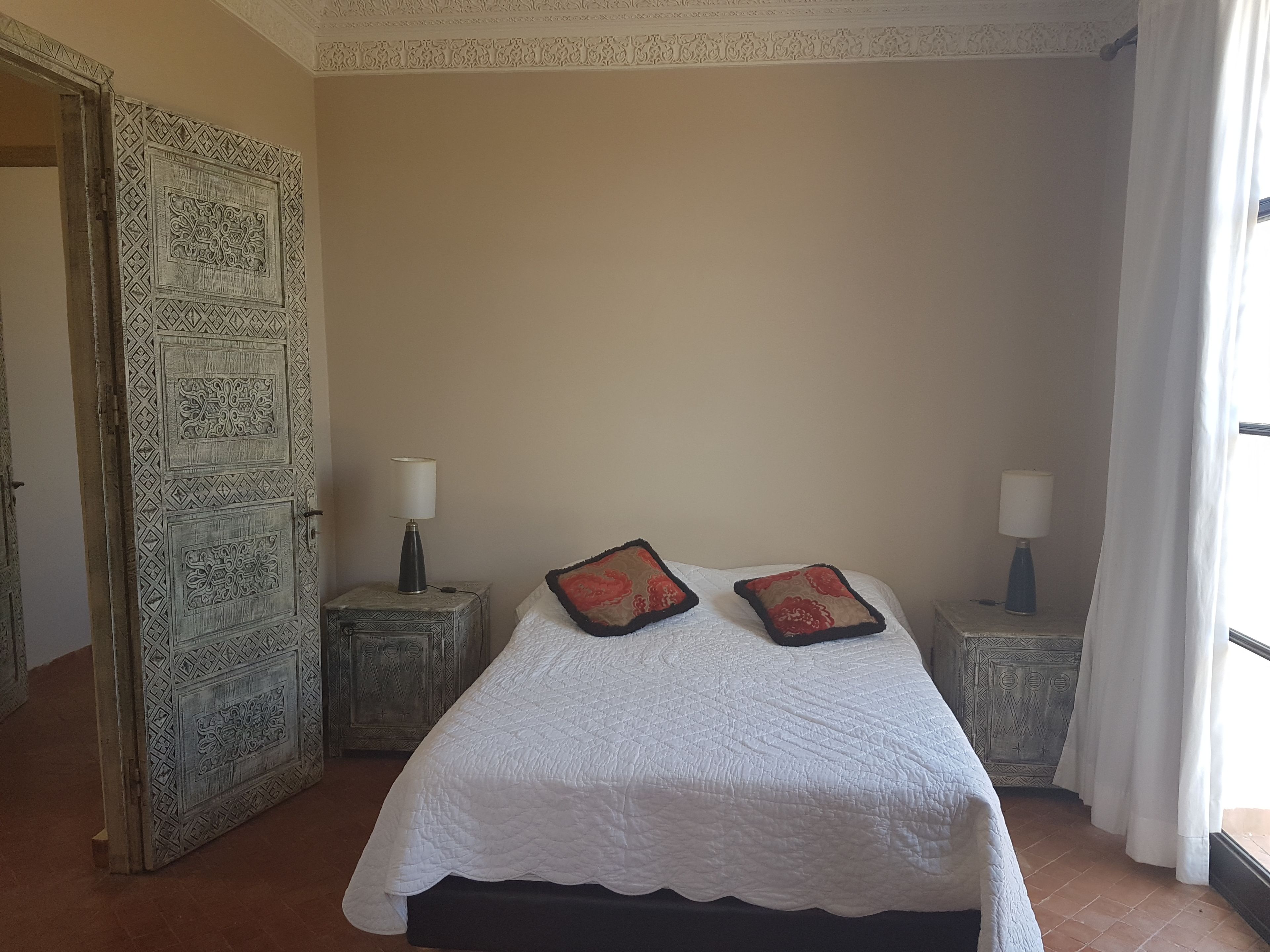 A bedroom with a bed, two nightstands, a decorative door, and a window with curtains.