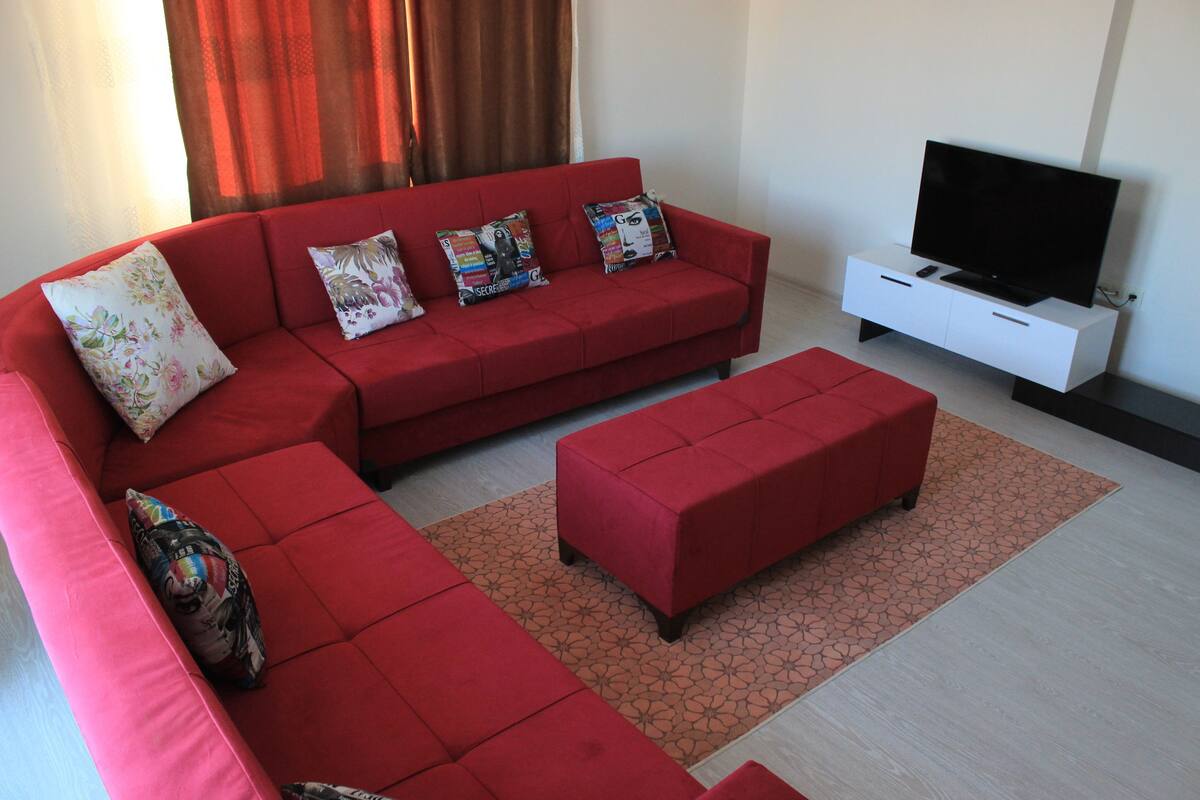 A modern living room with a red sectional sofa, matching ottoman, and a flat-screen TV on a white stand.