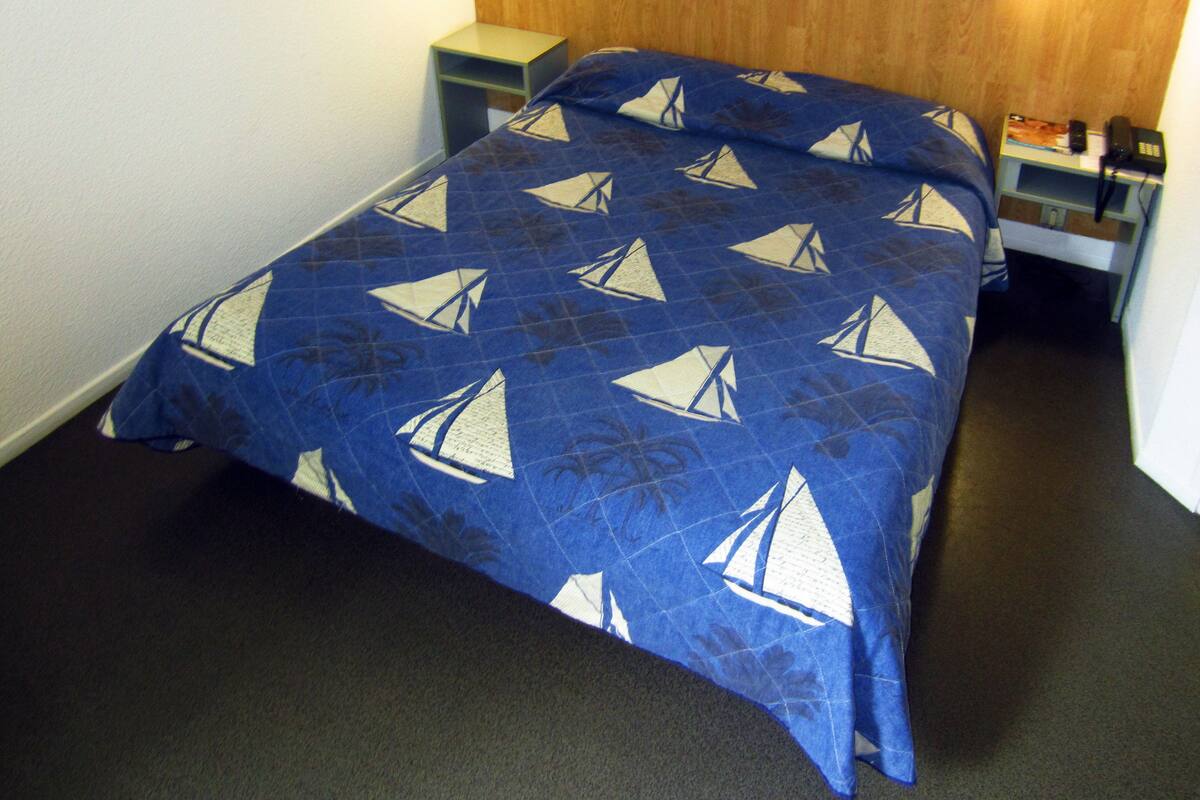 Cama com colcha estampada de veleiro azul e branco, cabeceira de madeira, duas mesas de cabeceira e um criado-mudo com abajur.