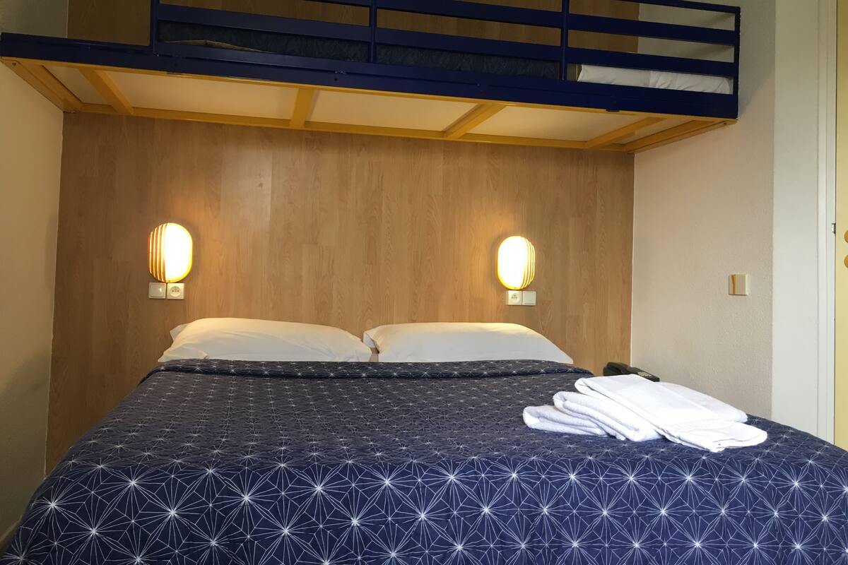 Uma cama beliche com cabeceira de madeira, duas luminárias de mesa e uma cama bem arrumada com colcha de padrão geométrico.