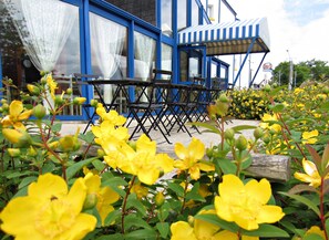 Un bâtiment bleu avec de grandes fenêtres et un auvent rayé. Il y a des tables et des chaises à l’extérieur.