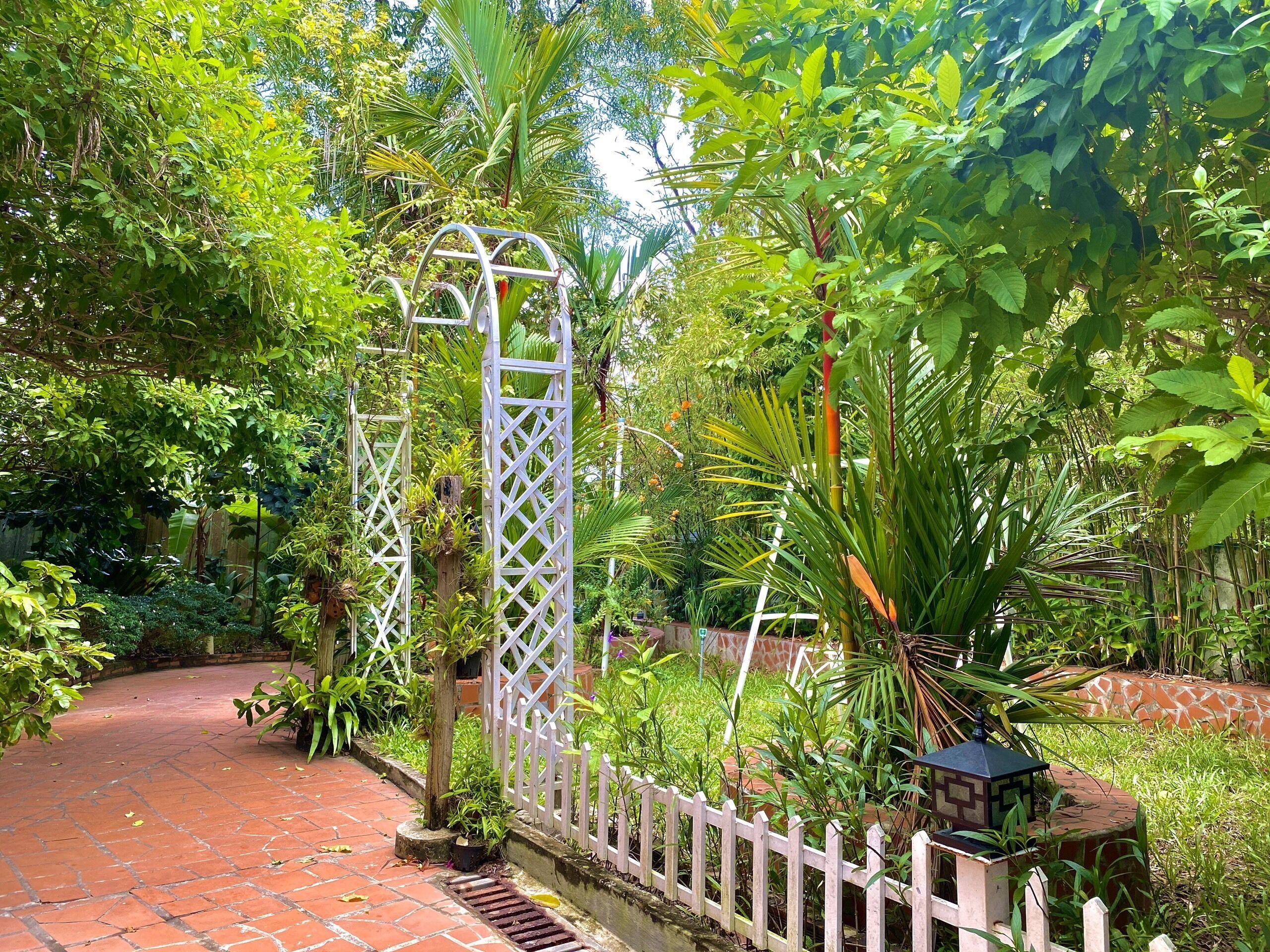 A garden pathway with a white lattice gate, surrounded by lush greenery and a brick wall.