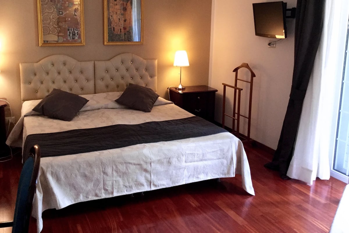 Habitación de hotel con una cama grande, dos cuadros en la pared, una mesita de noche con lámpara, un televisor montado y una ventana con cortinas.
