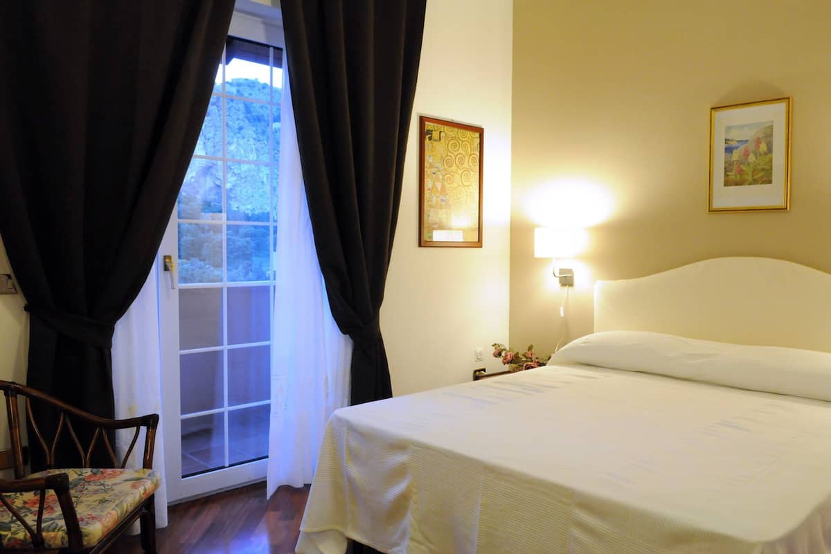 Una habitación de hotel con cama, una silla, ventana con cortinas y una lámpara fijada a la pared.