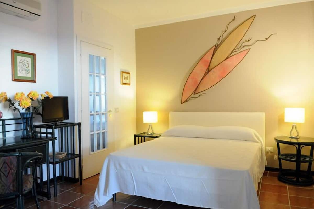Habitación de hotel con una cama, dos lámparas de noche, un escritorio con una silla, un televisor y un florero con flores.