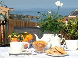 Una mesa dispuesta con alimentos para el desayuno, incluyendo un tazón de cereales, un plato de pasteles y una taza, con una vista panorámica del mar y las colinas al fondo.