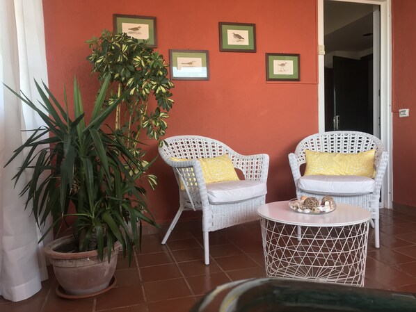 Una habitación con dos sillones blancos de mimbre con cojines amarillos, una mesa redonda blanca con un centro decorativo y una planta grande en maceta.