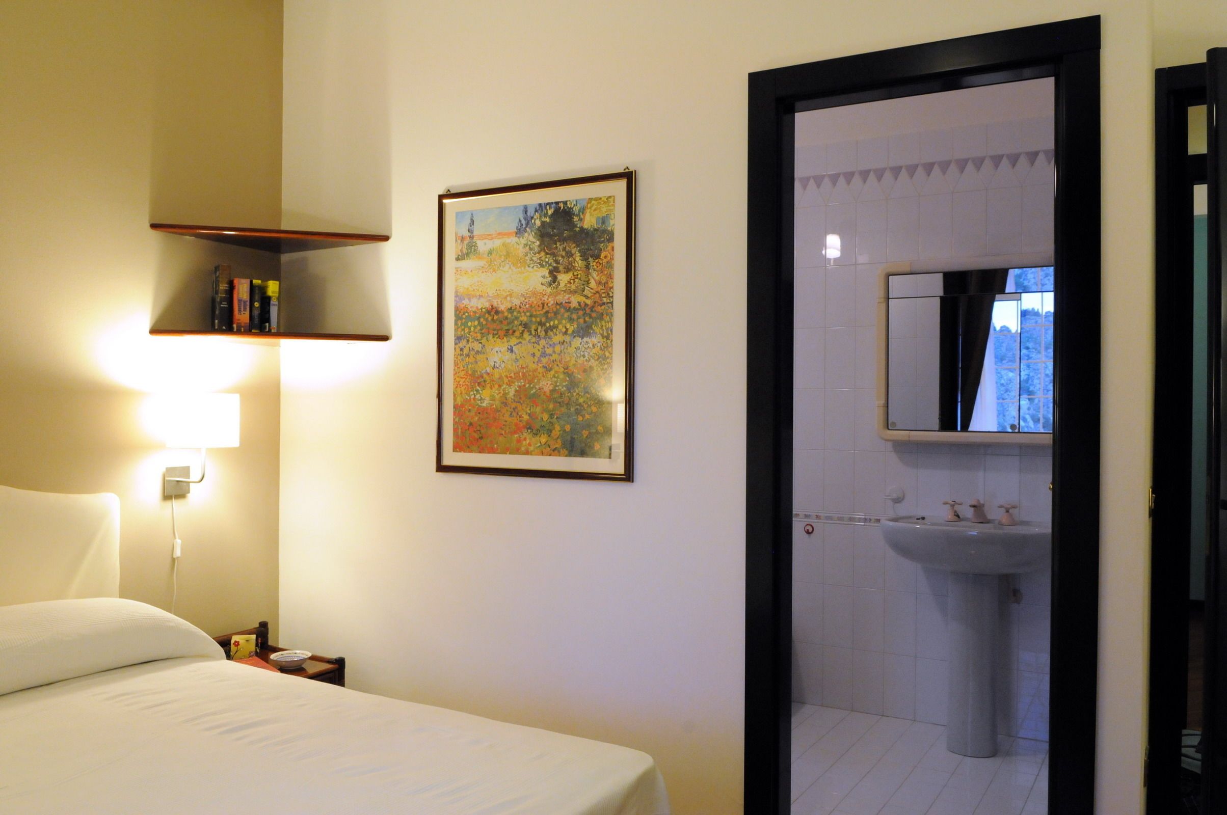 Una habitación de hotel con una cama, una mesita de noche, un estante con libros, un cuadro enmarcado y un baño visible a través de una puerta abierta.